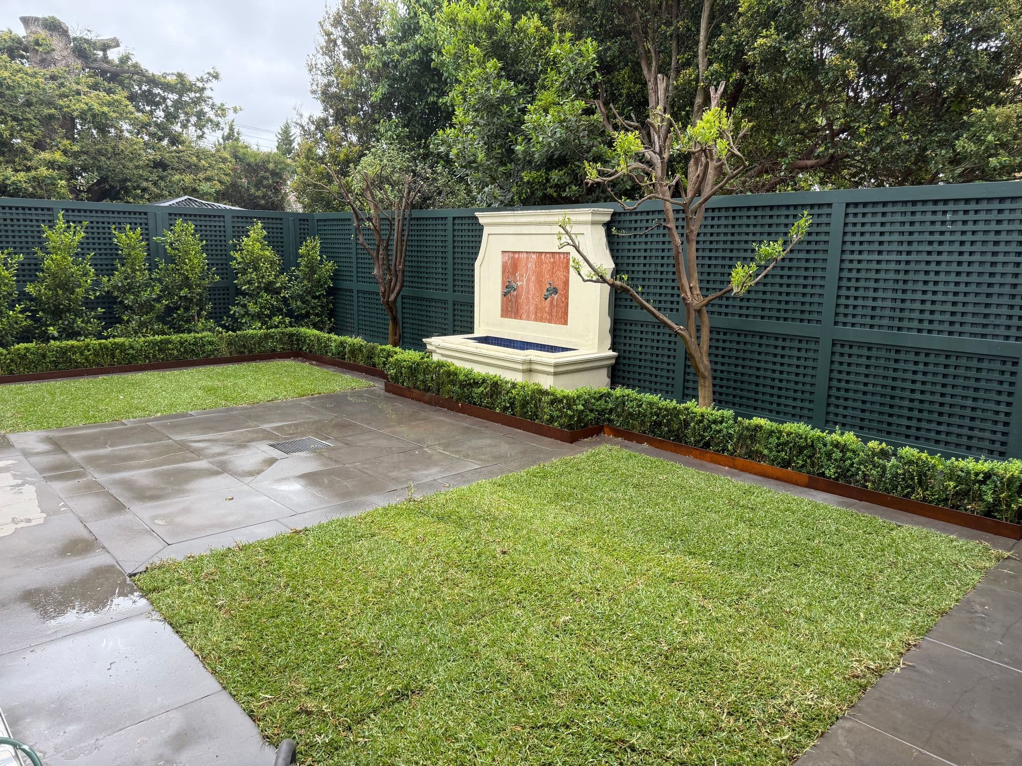 A new home's courtyard with fresh turf, stone paving, buxus hedges in corten edging and a wall fountain