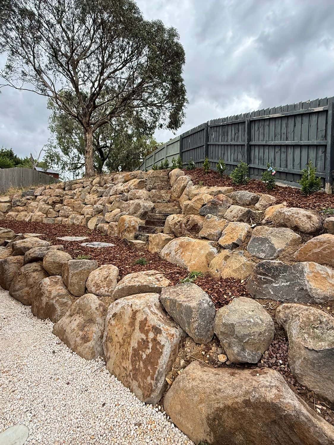 Terraced natural rock retaining walls with mulched planting beds along a boundary fence