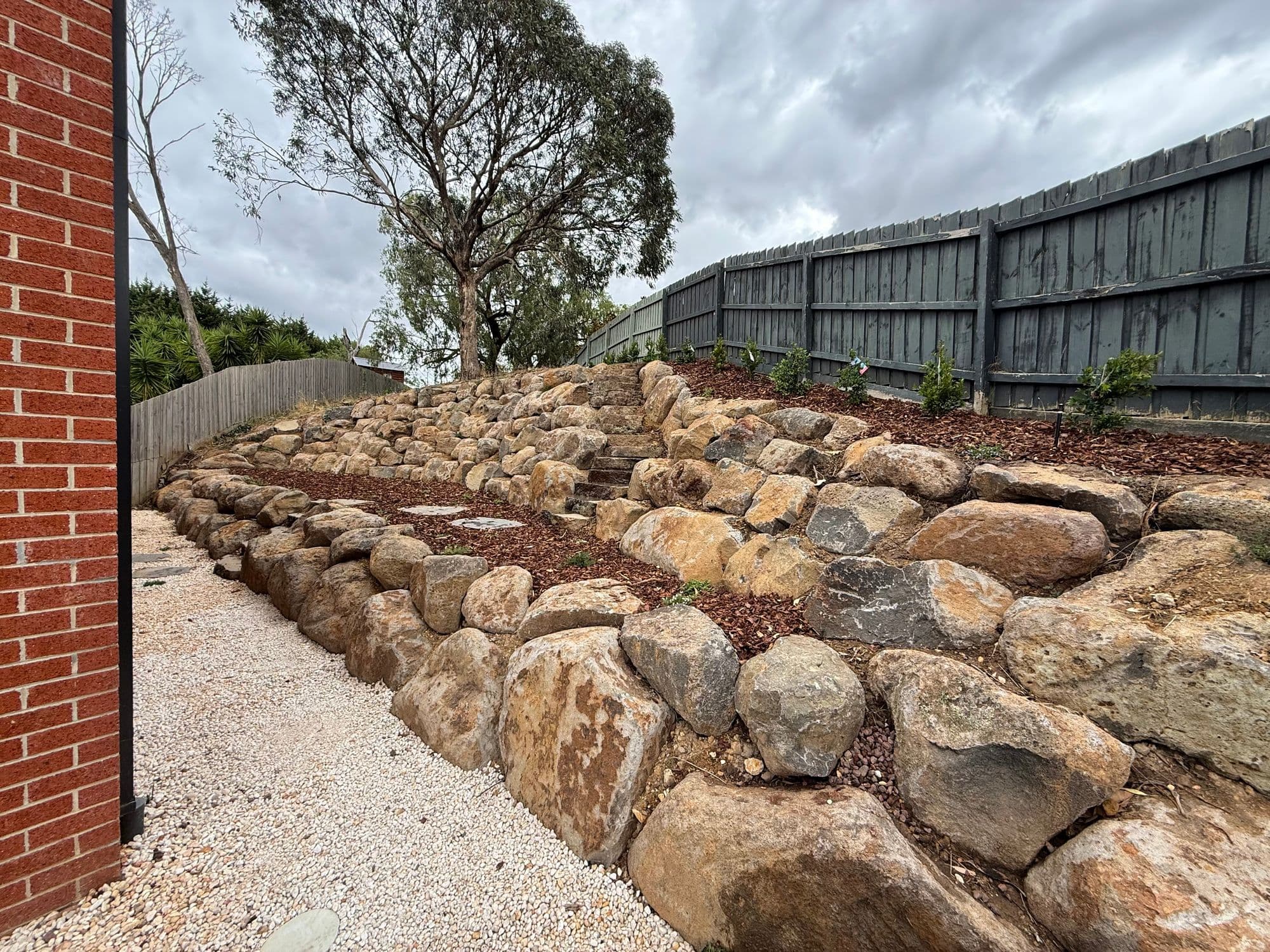 Terraced natural rock retaining walls with mulched planting beds along a boundary fence